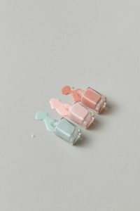Trois flacons de vernis à ongles pastel aux nuances rose pâle, pêche et bleu renversés avec art sur une surface grise douce.