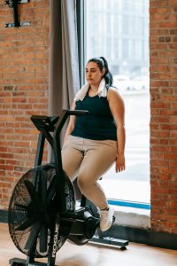 Full body of exhausted female sitting on exercise bike and putting arms down in gym after intense training Corps entier d'une femme épuisée assise sur un vélo d'exercice et baissant les bras en salle de sport après un entraînement intense