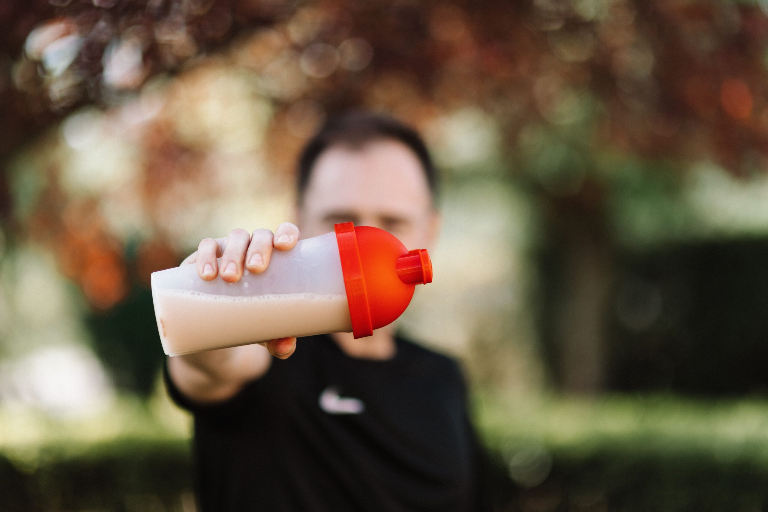 Focus on a man holding a plastic protein shake tumbler outdoors with a blurred background. Un homme tenant un verre de protéines en plastique à l'extérieur avec un arrière-plan flou.