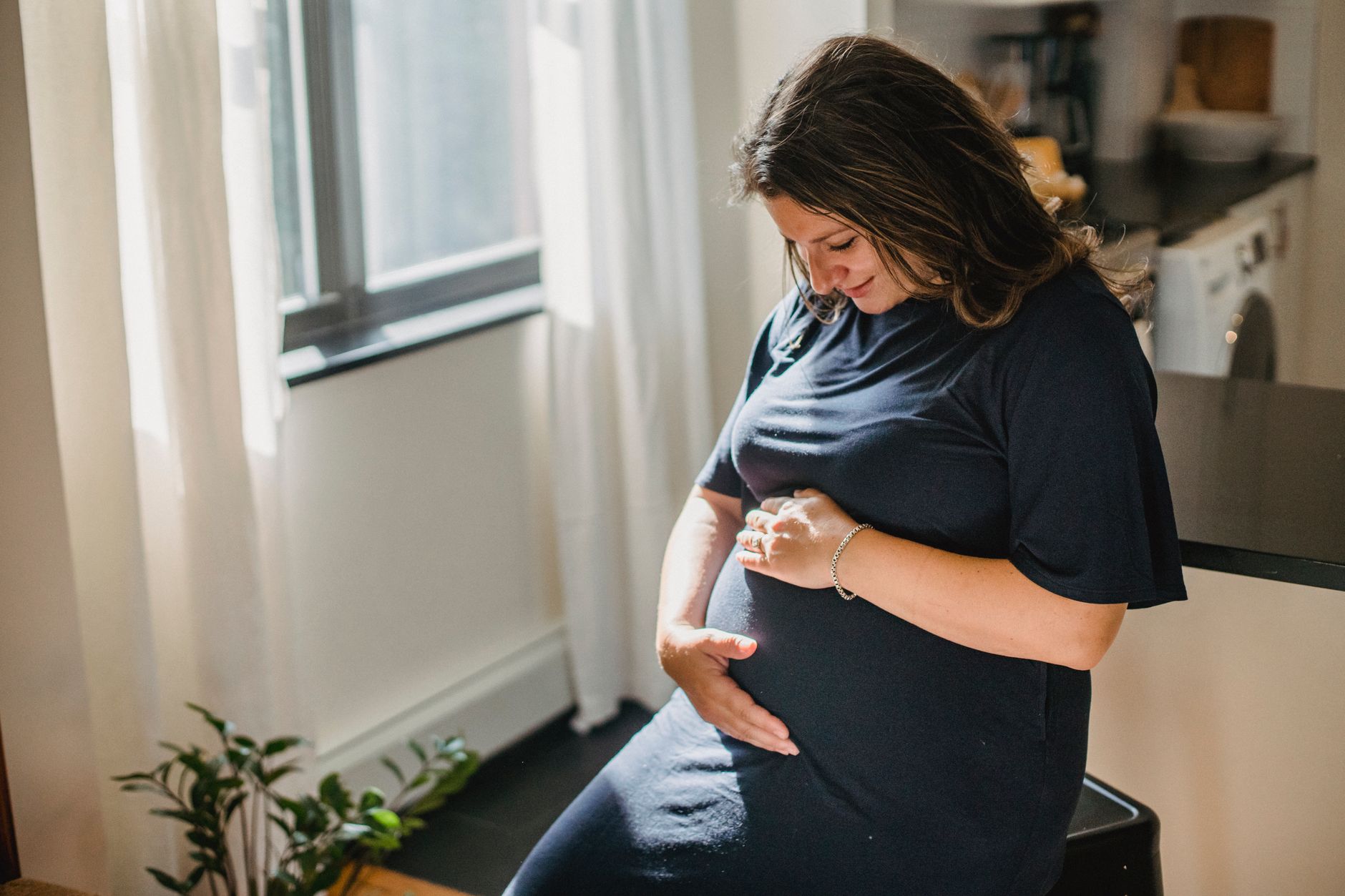 Femme enceinte assise, la main sur le ventre, dans un appartement lumineux